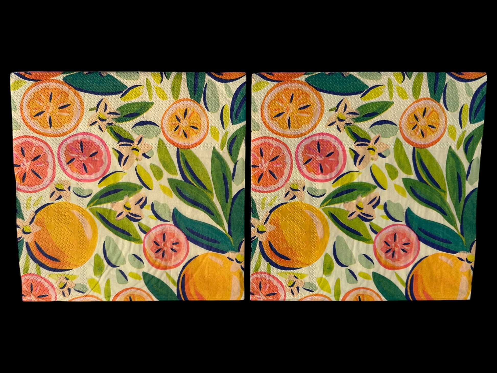 Close-up of two bright luncheon napkins featuring watercolor oranges, grapefruit, and green leaves designed by Kristen Vogel on a light cream background.