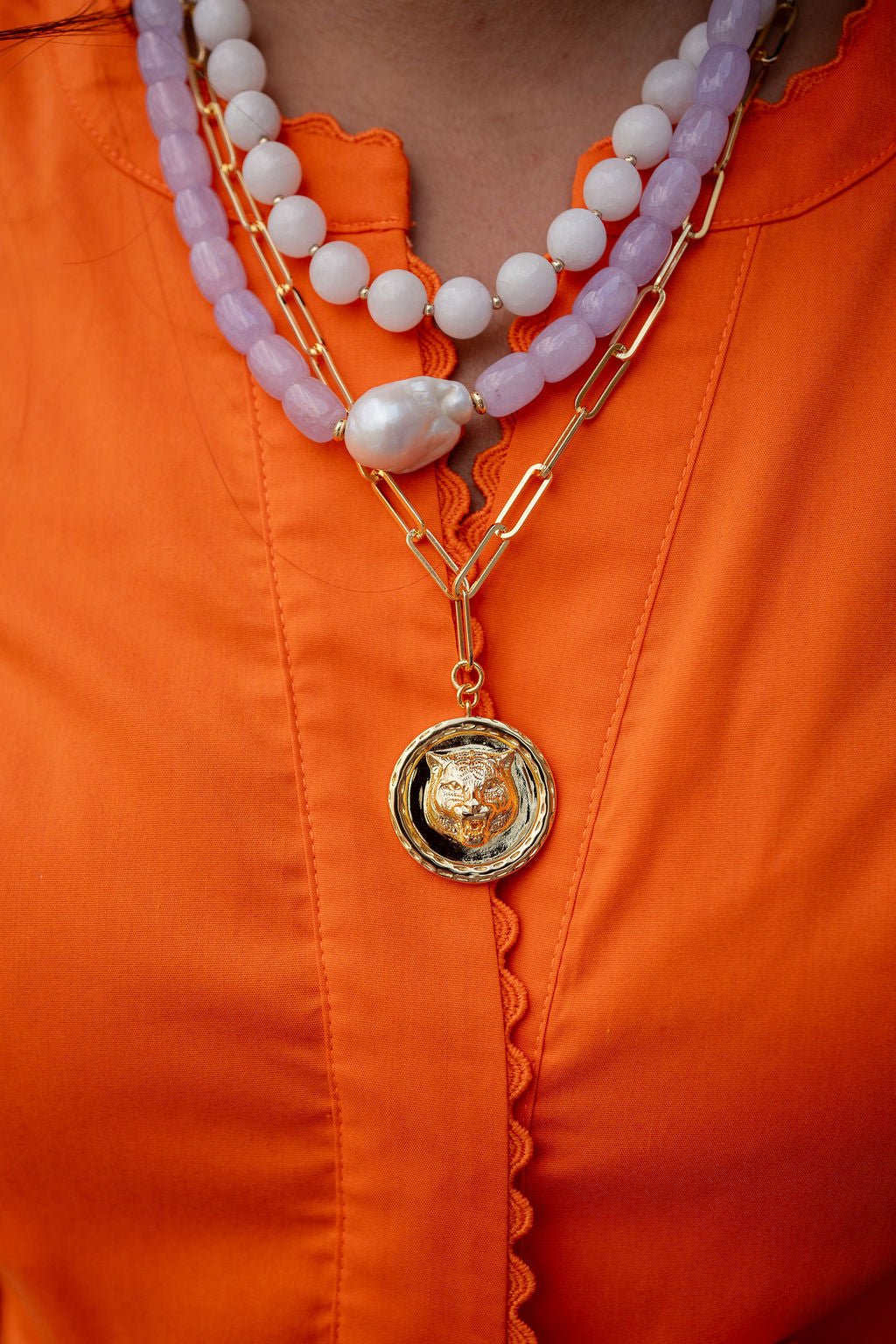 A close-up of a person wearing a layered necklace set over an orange blouse. The necklace features a gold chain with a circular pendant containing a gold tiger head, layered with a strand of white pearls and a lavender purple beaded strand.