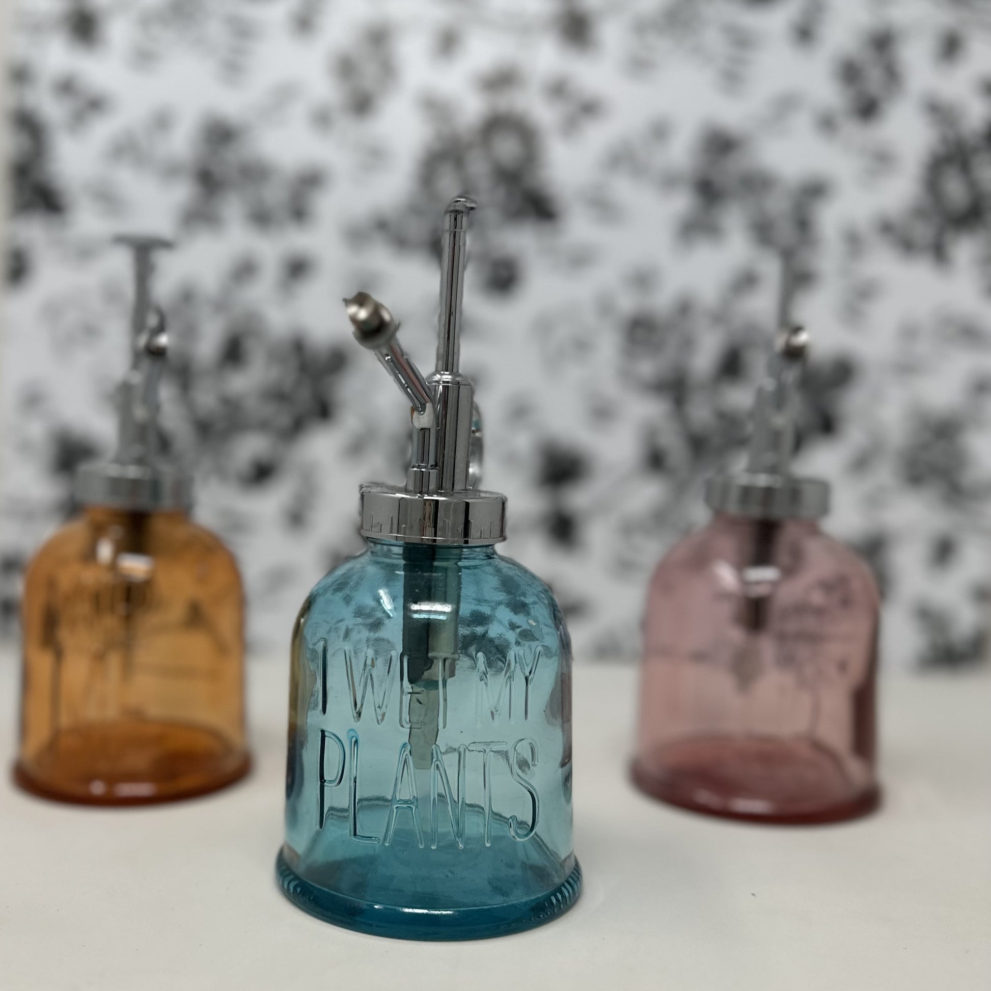 Three Mud Pie tinted glass plant misters in amber, blue, and pink with embossed sayings Water Me, I Wet My Plants, and Grow Up, with chrome pump sprayers, 6.25 x 3.5 inches