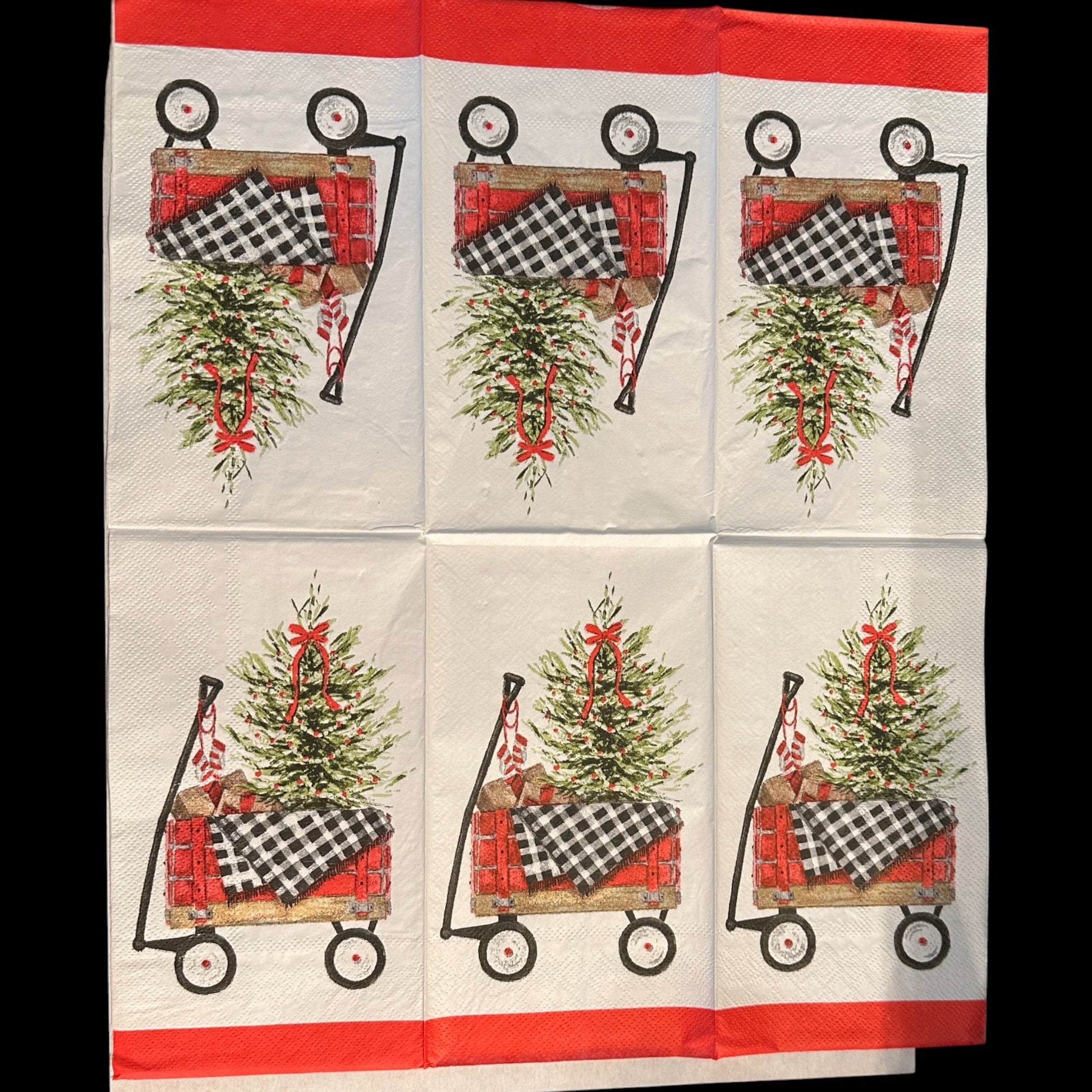 Close-up of an unfolded guest-size Christmas napkin featuring a red wagon carrying a decorated Christmas tree, wrapped gifts, and a black-and-white buffalo check blanket on a white background with a red border.
