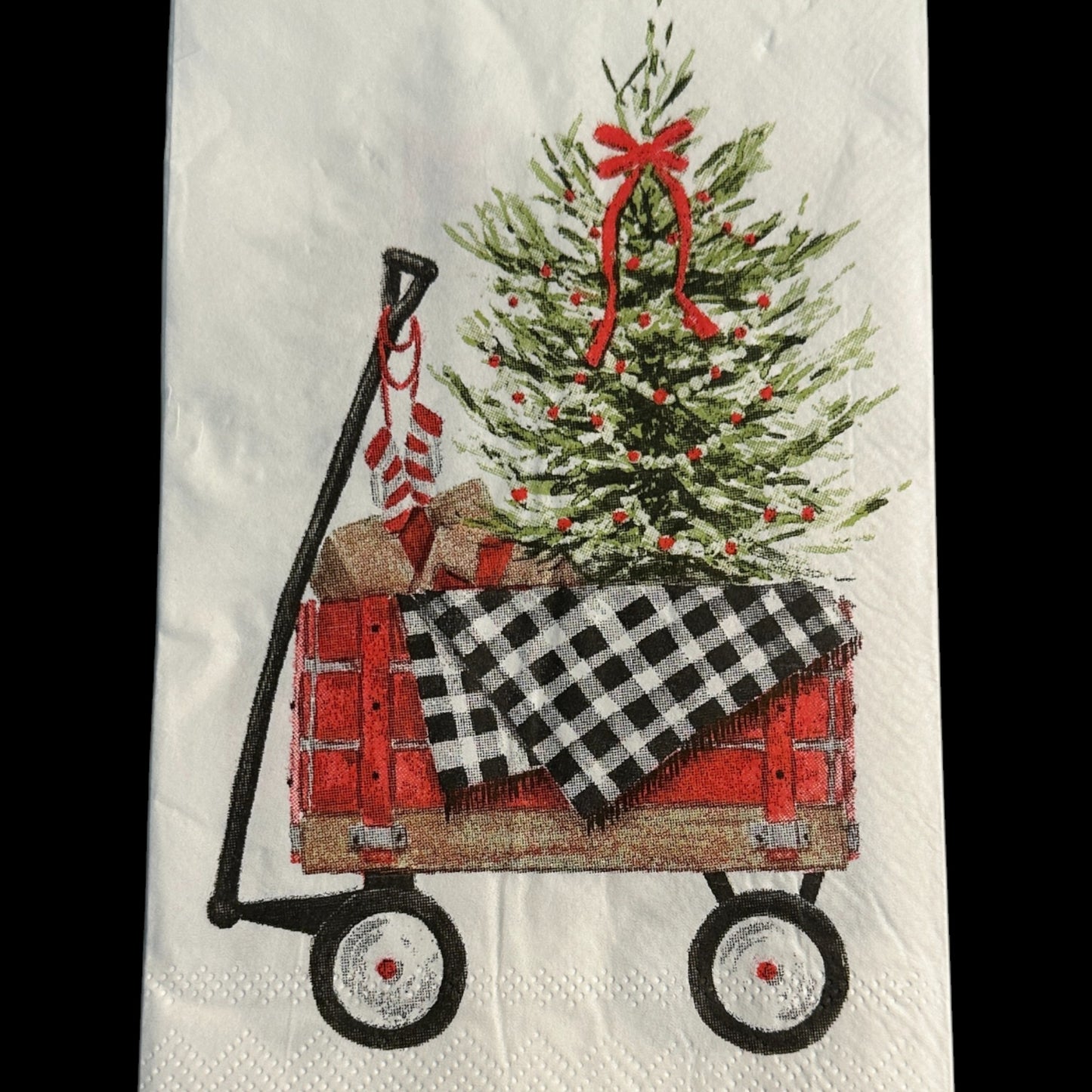 Close-up of a guest-size Christmas napkin featuring a red wagon carrying a decorated Christmas tree, wrapped gifts, and a black-and-white buffalo check blanket on a white background with a red border.
