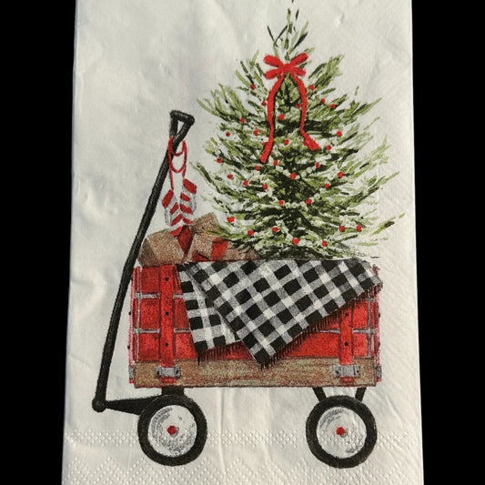 Close-up of a guest-size Christmas napkin featuring a red wagon carrying a decorated Christmas tree, wrapped gifts, and a black-and-white buffalo check blanket on a white background with a red border.
